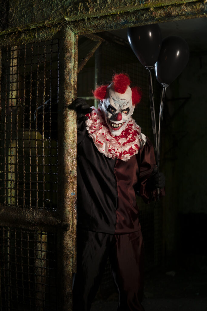 scary clown with red hair and a sinister smile on his face standing in a haunted house during Halloween event. 