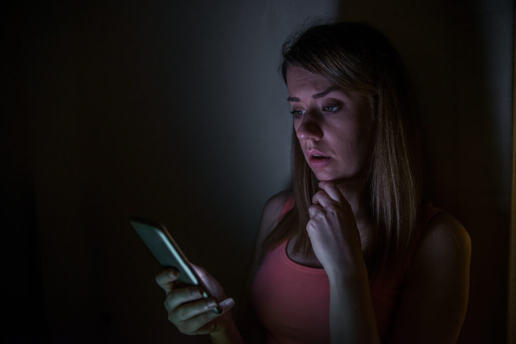 A woman in a dark room looking at her phone in distress.