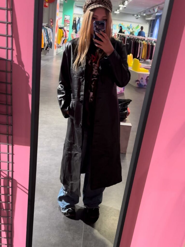 A woman taking a selfie in a mirror with a long leather coat on. 